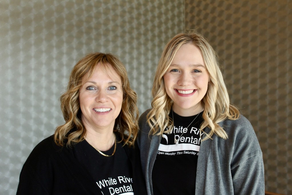 Two smiling women, one in a black shirt and the other in a grey cardigan over a black clinic shirt, posing together against a neutral background.