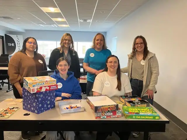 Five women gathered around a table filled with board games and toys, smiling for a group photo in an office setting.