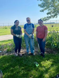 Three women standing together in a grassy outdoor area near a garden during a community project.