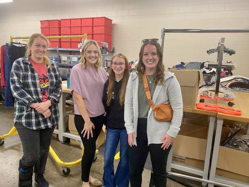women smiling and posing together in a storage room filled with organized supplies and donation boxes.