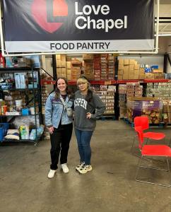 Two women posing together in front of a Love Chapel Food Market sign during a community outreach event