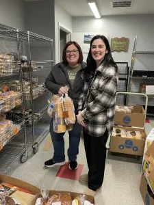 Two women smiling and holding grocery bags while volunteering in a well-stocked food pantry