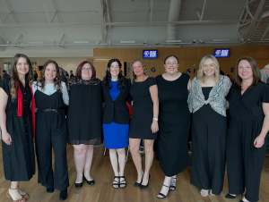A group of women in professional black and blue attire posing together for a formal group photo in an indoor venue