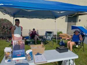 Three women participating in an outdoor community event under a blue canopy tent, featuring a table with brochures, a donation basket, and sign-up sheets.