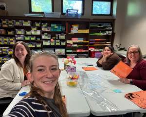 A group of four women smiling and working together at a large white table in a room filled with organized shelves of supplies.